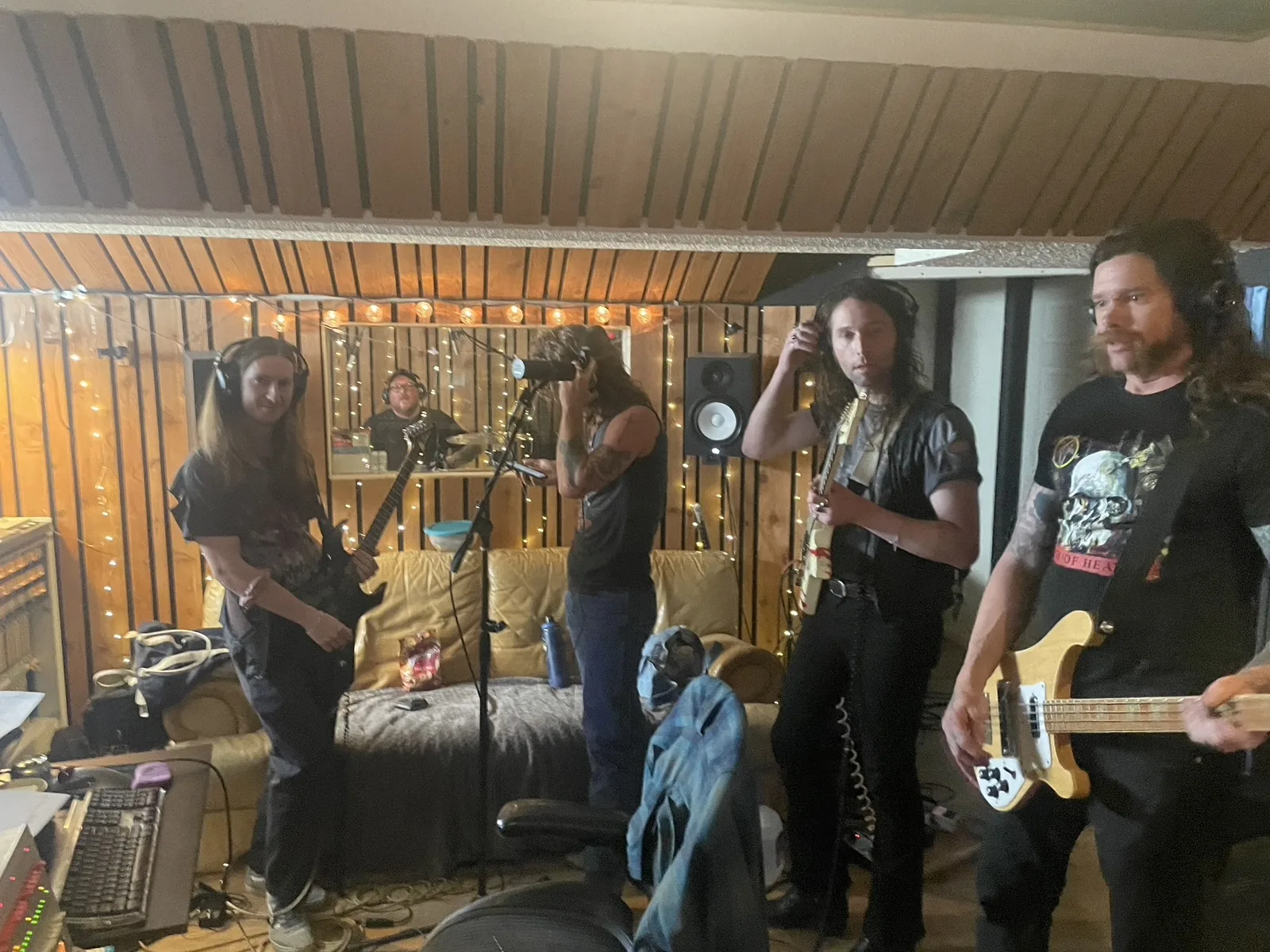 Musicians with guitars, bass and headphones, with a drummer visible through the studio window
