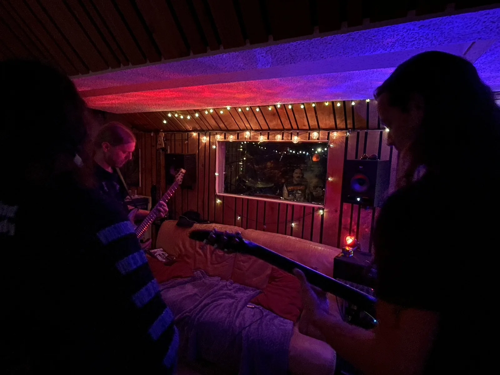 Musicians playing guitars with a drummer visible through the studio window